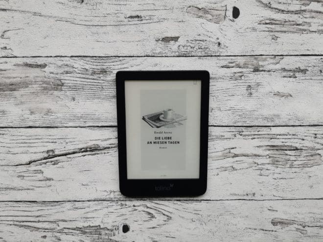 Das eBook "Die Liebe an miesen Tagen" von Ewald Arenz auf einem Tolino, der auf einer hellen, holzähnlichen Oberfläche liegt ist auf dem Bild. Das Bild ist das Headerbild zur Rezension des Buches.