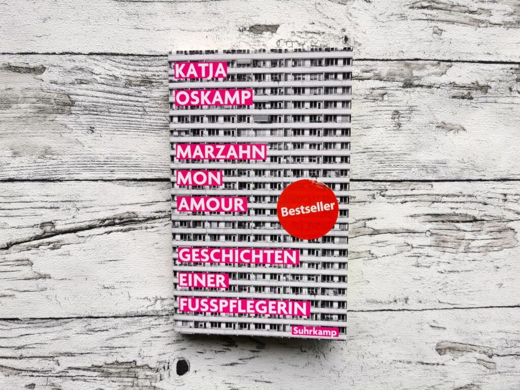Auf dem Bild ist das Buch Marzahn Mon Amour von Katja Oskamp auf einem hellen, holzähnlichen Hintergrund zu sehen. Das Bild ist das Headerbild des Beitrags. Der Beitrag ist eine Rezension des Buches.