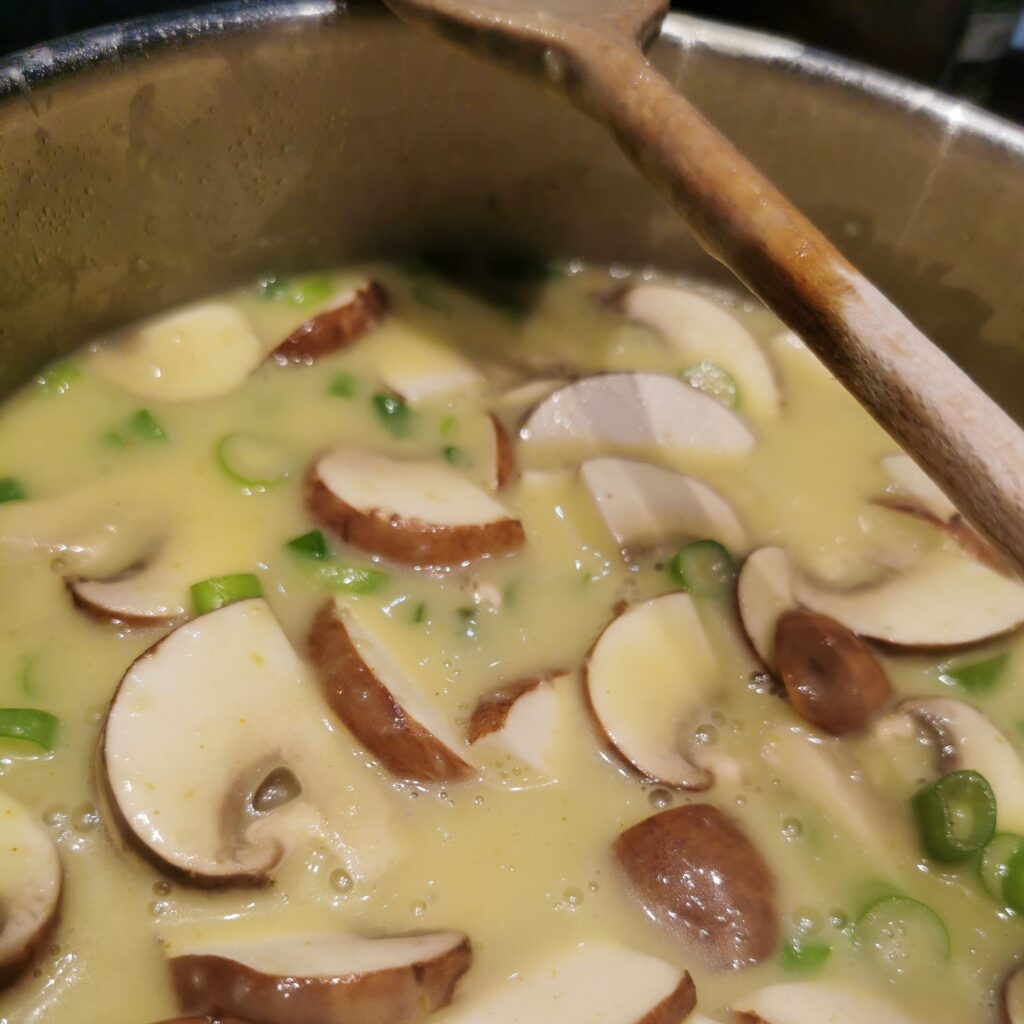 Suppe mit Frühlingszwiebeln, Kartoffeln, Ingwer und Champignons in einem Edelstahlkochtopf. Rechts liegt ein Holzkochlöffel auf dem Rand des Topfes.