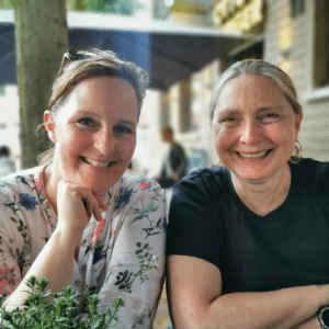 Auf dem Foto sind Valerie Wagner und Antje Tomfohrde abgebildet. Sie sitzen nebeneinander an einem Tisch und lächeln in die Kamera.