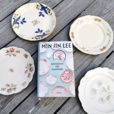 Auf dem Foto ist das Buch Gratisessen für Millionäre von Min Jin Lee auf einem Holztisch liegend, von vier Tellern umgeben abgebildet. Es ist das Headerbild zur Rezension des Buchs.