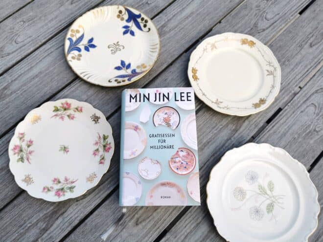Auf dem Foto ist das Buch Gratisessen für Millionäre von Min Jin Lee auf einem Holztisch liegend, von vier Tellern umgeben abgebildet. Es ist das Headerbild zur Rezension des Buchs.