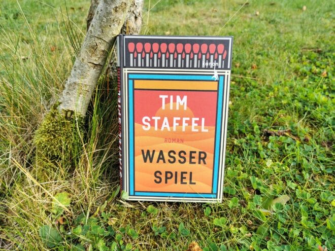 Auf dem Foto ist das Buch Wasserspiel von Tim Staffel an einen Baumstamm gelehnt auf einer Wiese fotografiert. Es ist das Header-Bild zur Rezension des Buchs.