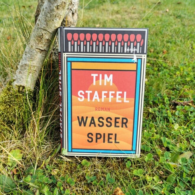 Auf dem Foto ist das Buch Wasserspiel von Tim Staffel an einen Baumstamm gelehnt auf einer Wiese fotografiert. Es ist das Header-Bild zur Rezension des Buchs.