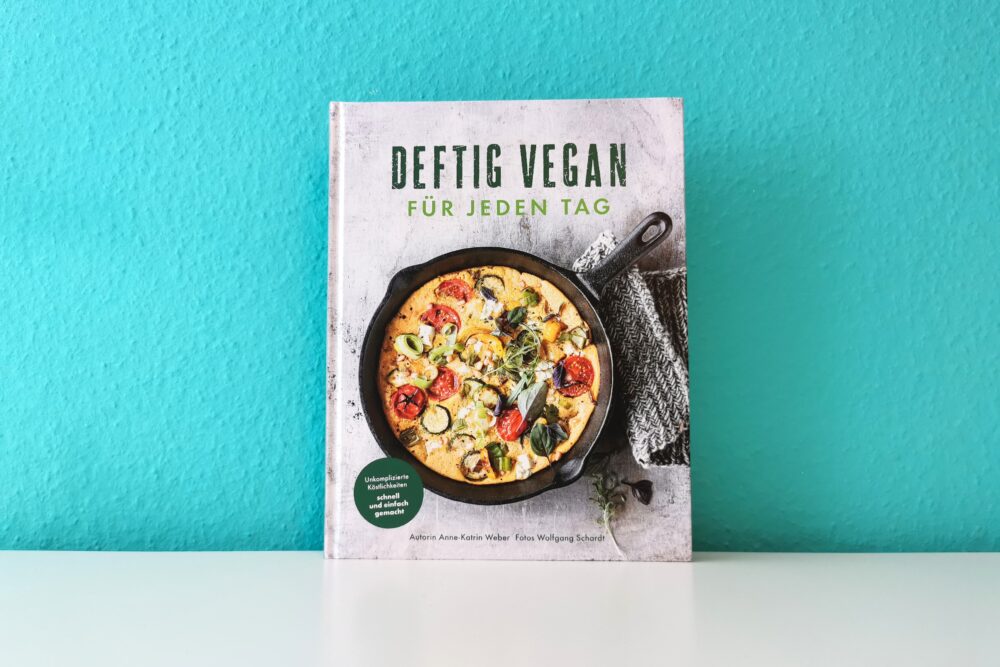 Auf dem Foto ist das Buch Deftig vegan für jeden Tag von Anne-Katrin Weber vor einer türkisen Wand auf einer weißen Oberfläche fotografiert. Es ist das Headerbild zur Rezension des Buches.