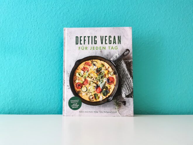 Auf dem Foto ist das Buch Deftig vegan für jeden Tag von Anne-Katrin Weber vor einer türkisen Wand auf einer weißen Oberfläche fotografiert. Es ist das Headerbild zur Rezension des Buches.