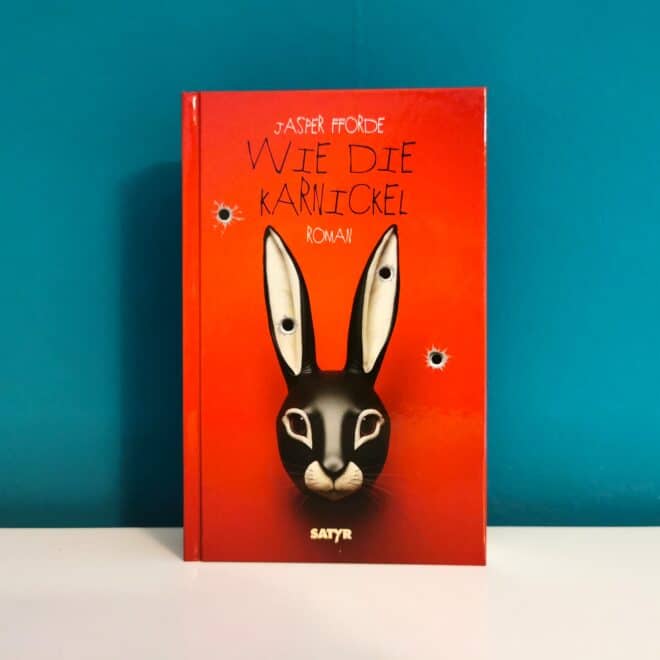 Auf dem Foto ist das Buch Wie die Karnickel von Jasper Fforde vor einer dunkelgrünen Wand auf einer glatten weißen Fläche stehend abgebildet. Es ist das Header-Bild meiner Rezension des Buchs.