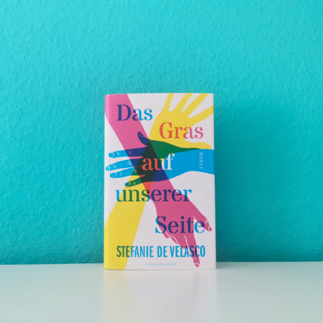 Auf dem Foto ist das Buch Das Gras auf unserer Seite von Stefanie de Velasco vor einer türkisen Wand auf einer glatten weißen Fläche fotografiert. Es ist das Headerbild der dazugehörigen Rezension.