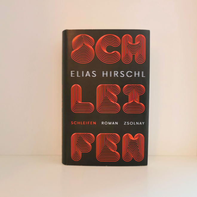 Auf dem Foto ist das Buch Schleifen von Elias Hirschl in einem Regalfach eines weißen Billy Regals fotografiert worden. Das Bild ist das Headerbild zur Rezension des Buchs.