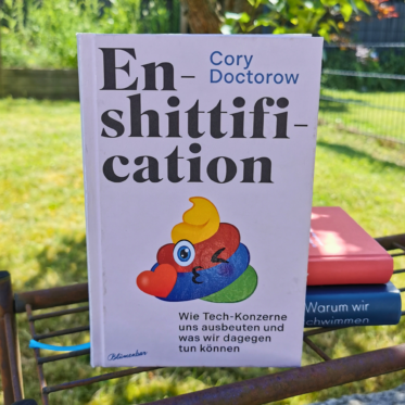 Das Buch "Enshittification" von Cory Doctorow (deutsche Ausgabe, Blumenbar) steht aufgeschlagen auf einem rostigen Metallgitter im Garten, im Hintergrund grüner Rasen und ein Zaun. Auf dem cremefarbenen Cover ist eine bunte, augenzwinkernde Kot-Emoji-Figur mit Herz abgebildet. Im Hintergrund liegen zwei weitere Bücher gestapelt. Es ist das Header-Bild zu einer Rezension des Buchs.