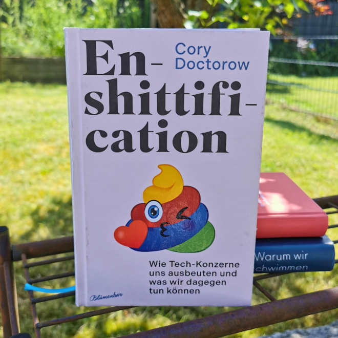 Das Buch "Enshittification" von Cory Doctorow (deutsche Ausgabe, Blumenbar) steht aufgeschlagen auf einem rostigen Metallgitter im Garten, im Hintergrund grüner Rasen und ein Zaun. Auf dem cremefarbenen Cover ist eine bunte, augenzwinkernde Kot-Emoji-Figur mit Herz abgebildet. Im Hintergrund liegen zwei weitere Bücher gestapelt. Es ist das Header-Bild zu einer Rezension des Buchs.