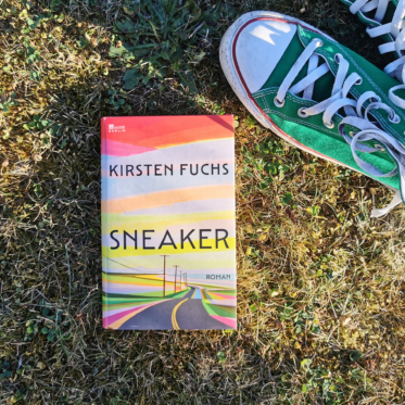 Das Buch „Sneaker" von Kirsten Fuchs liegt aufrecht im sonnigen, teils vertrockneten Gras. Das Cover zeigt eine in bunten Streifen gemalte Landschaft mit einer Straße, die zwischen Strommasten in den Horizont führt. Rechts daneben steht ein getragener grüner Sneaker (Converse Chuck) mit weißer Kappe und heller Schnürung. Bildtext: rowohlt Berlin. Kirsten Fuchs. Sneaker. Roman.