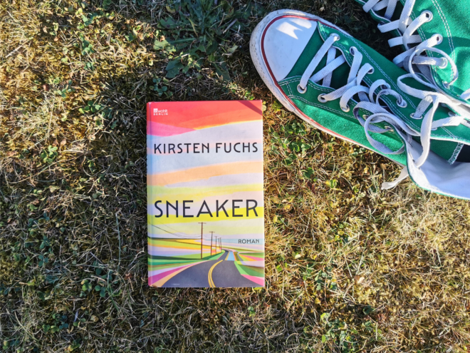 Das Buch „Sneaker" von Kirsten Fuchs liegt aufrecht im sonnigen, teils vertrockneten Gras. Das Cover zeigt eine in bunten Streifen gemalte Landschaft mit einer Straße, die zwischen Strommasten in den Horizont führt. Rechts daneben steht ein getragener grüner Sneaker (Converse Chuck) mit weißer Kappe und heller Schnürung. Bildtext: rowohlt Berlin. Kirsten Fuchs. Sneaker. Roman.
