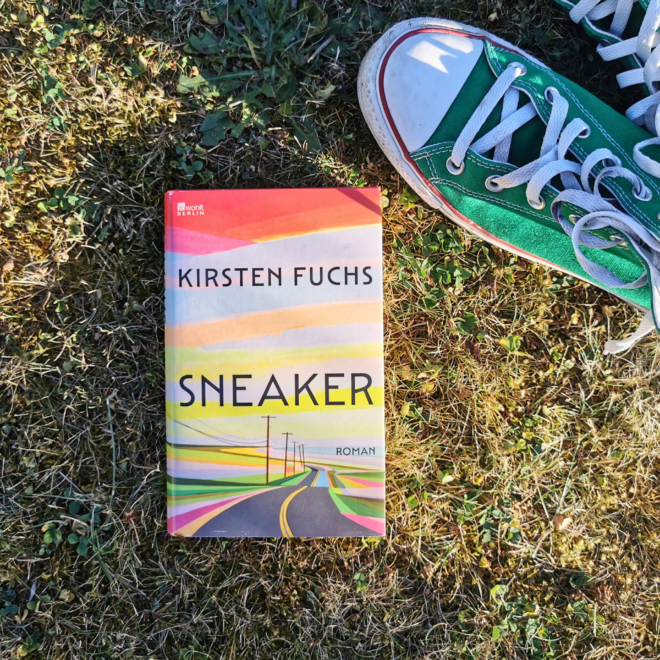 Das Buch „Sneaker" von Kirsten Fuchs liegt aufrecht im sonnigen, teils vertrockneten Gras. Das Cover zeigt eine in bunten Streifen gemalte Landschaft mit einer Straße, die zwischen Strommasten in den Horizont führt. Rechts daneben steht ein getragener grüner Sneaker (Converse Chuck) mit weißer Kappe und heller Schnürung. Bildtext: rowohlt Berlin. Kirsten Fuchs. Sneaker. Roman.