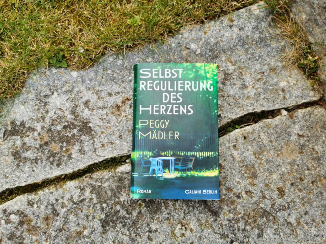 Das Buch Selbstregulierung des Herzens von Peggy Mädler, erschienen bei Galiani Berlin, liegt aufrecht auf einer verwitterten Steinplatte, an deren oberem Rand Gras und Moos wachsen. Das Cover zeigt in Grün- und Blautönen einen dämmrigen Garten mit Tisch und Stühlen vor einem Zaun; Titel in Weiß, Autorinnenname in Gelb.