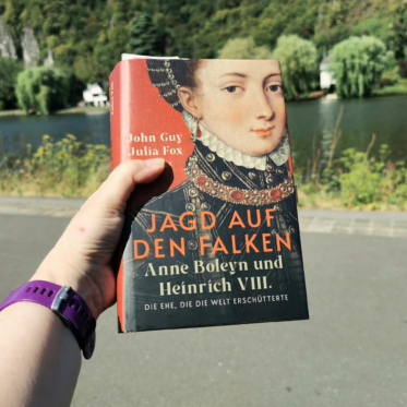 Eine Hand mit lila Sportuhr am Handgelenk hält das gebundene Buch „Jagd auf den Falken. Anne Boleyn und Heinrich VIII. Die Ehe, die die Welt erschütterte“ von John Guy und Julia Fox. Das Cover zeigt ein Renaissance-Porträt einer jungen Frau mit Perlenhaube, weißer Halskrause und juwelenbesetztem Kragen vor rotem Hintergrund. Im Hintergrund liegen ein Fluss, Weiden, ein weißes Haus und steile, bewaldete Felsen in sommerlichem Licht.