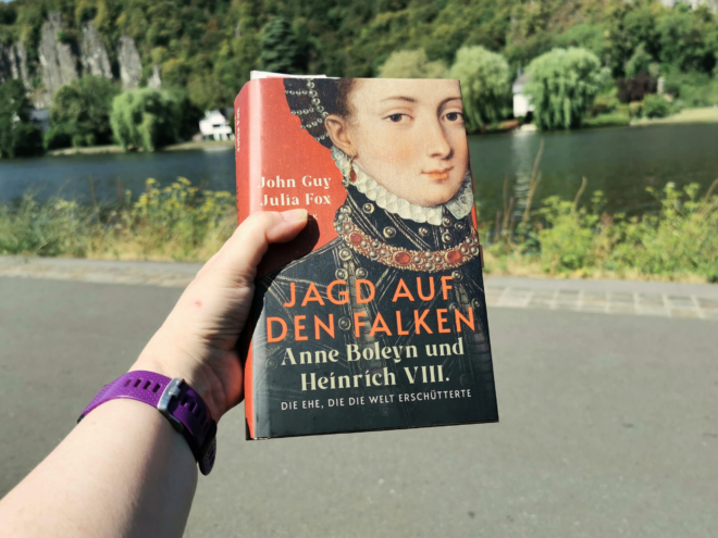 Eine Hand mit lila Sportuhr am Handgelenk hält das gebundene Buch „Jagd auf den Falken. Anne Boleyn und Heinrich VIII. Die Ehe, die die Welt erschütterte“ von John Guy und Julia Fox. Das Cover zeigt ein Renaissance-Porträt einer jungen Frau mit Perlenhaube, weißer Halskrause und juwelenbesetztem Kragen vor rotem Hintergrund. Im Hintergrund liegen ein Fluss, Weiden, ein weißes Haus und steile, bewaldete Felsen in sommerlichem Licht.