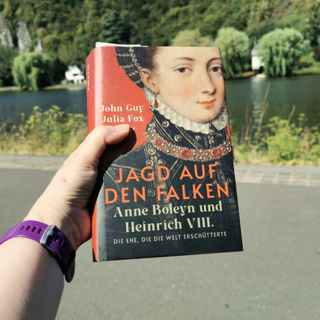 Eine Hand mit lila Sportuhr am Handgelenk hält das gebundene Buch „Jagd auf den Falken. Anne Boleyn und Heinrich VIII. Die Ehe, die die Welt erschütterte“ von John Guy und Julia Fox. Das Cover zeigt ein Renaissance-Porträt einer jungen Frau mit Perlenhaube, weißer Halskrause und juwelenbesetztem Kragen vor rotem Hintergrund. Im Hintergrund liegen ein Fluss, Weiden, ein weißes Haus und steile, bewaldete Felsen in sommerlichem Licht.